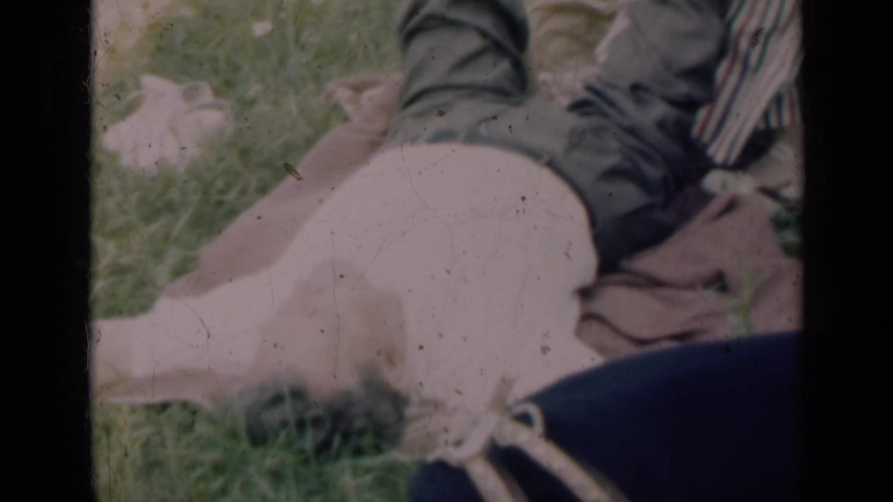 Faded 8mm frame from Indiana, 1948, with a man's bare legs extended on grass while a woman sits nearby.