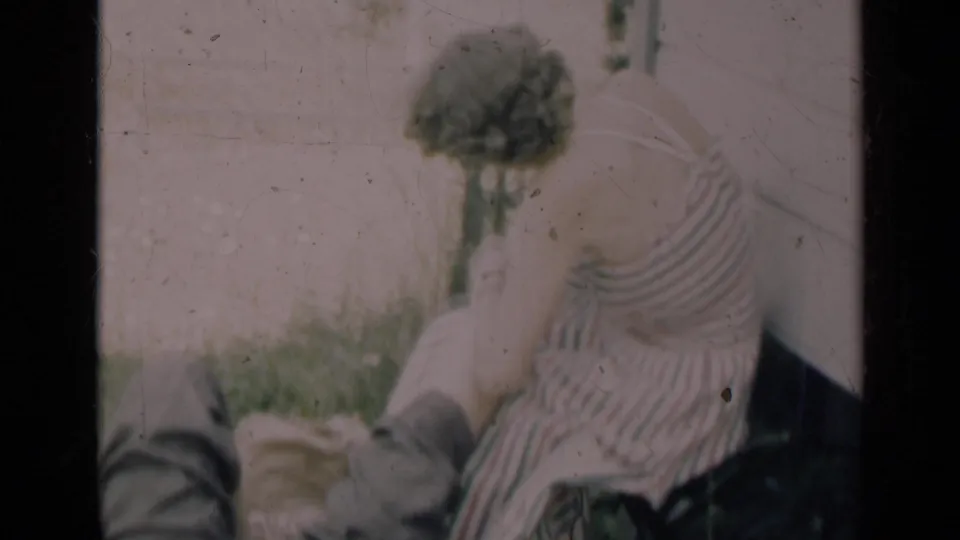 Soft 8mm frame from Indiana, 1948, with a woman bending over a man resting on grass near a pale exterior wall.