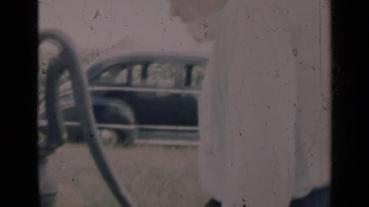 In Indiana 1948 home-movie footage, an adult stands near a hand water pump with a rounded vintage car behind them.