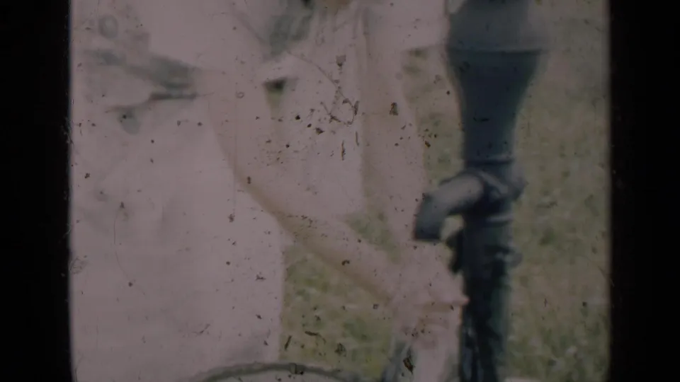 An adult stands beside the hand pump as the spout and handle dominate the foreground of the faded 8mm frame.