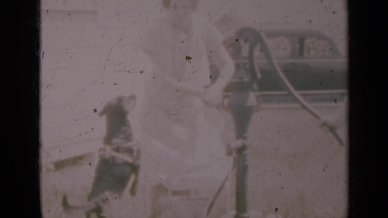 In 1948 Indiana home-movie footage, a woman stands by a hand pump while a dog is nearby and a vintage car sits in the background.