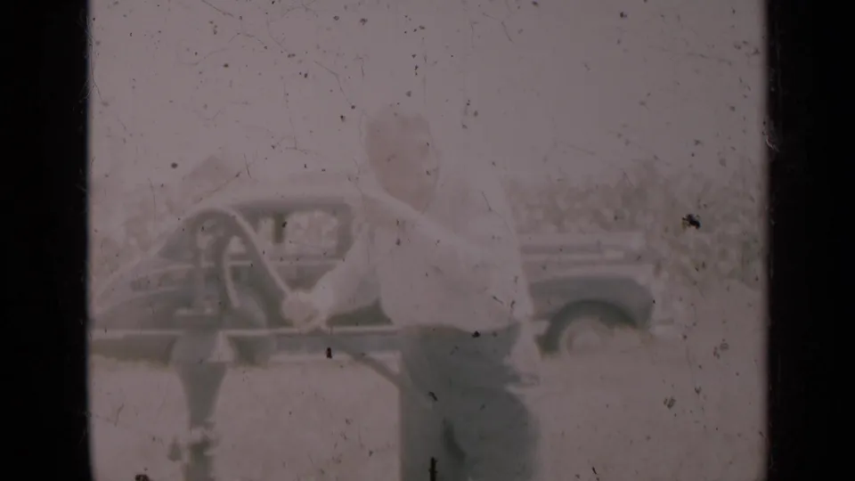 A woman leans near the hand pump in the Indiana scene, with the pump handle and vintage automobile visible through the faded 8mm frame.