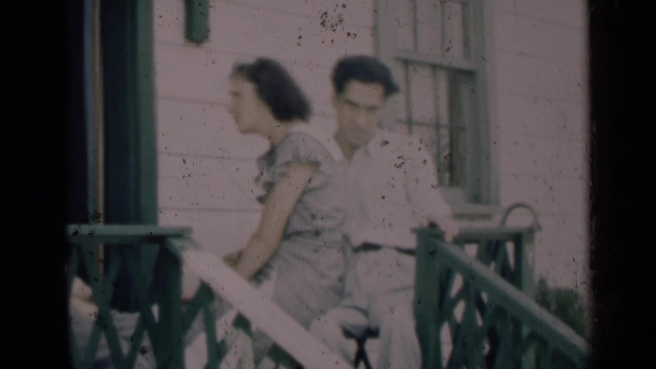 In 1948 Indiana home-movie footage, a young woman and man sit on a porch near green railings and house siding.