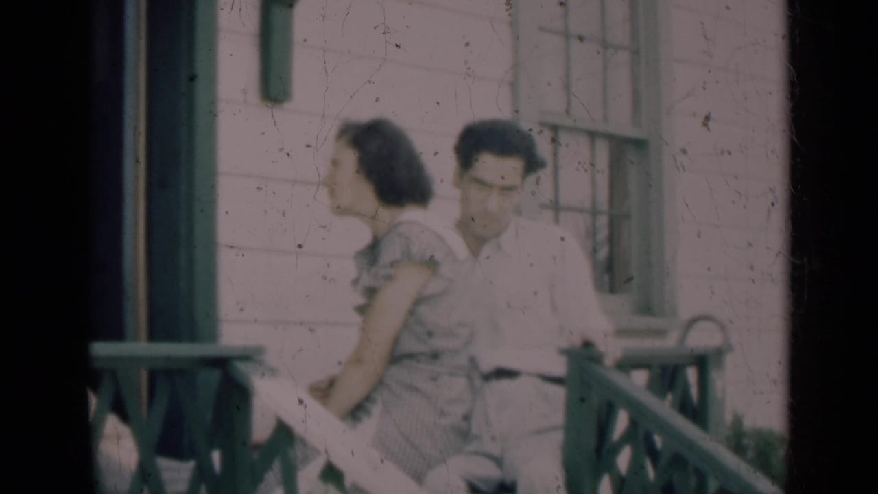 The young couple remains seated on the porch, partly framed by dark 8mm film edges and the house exterior.