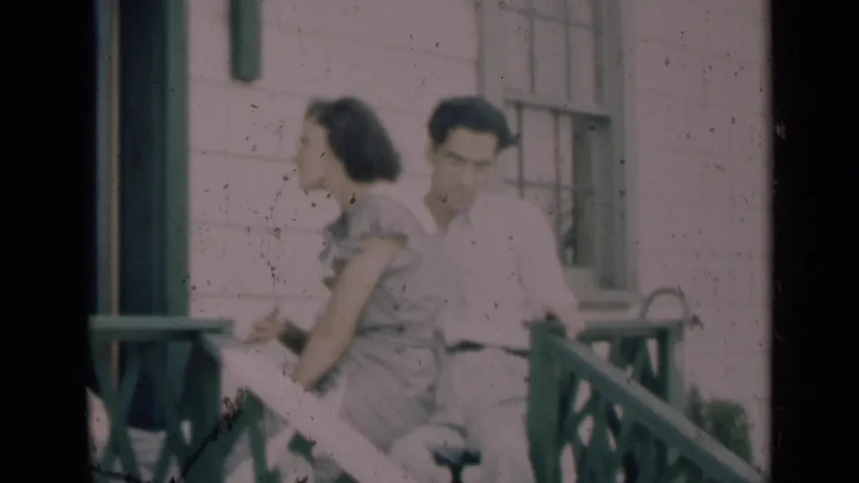 A young woman in a patterned dress and a man in a light shirt sit close together on the porch in soft 8mm color.