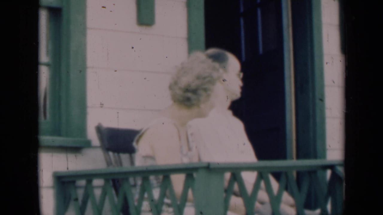 Older couple seated on an Indiana porch in 1948 home-movie footage, framed by green trim and pale clapboard siding.
