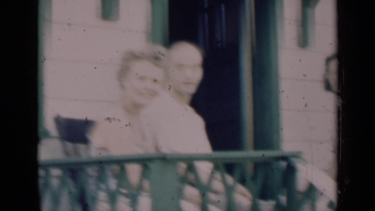 Soft 8mm frame of an older couple on a porch in Indiana, with the woman turned toward the camera and green trim behind them.