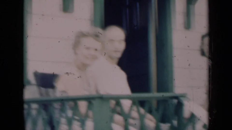 Soft 8mm frame of an older couple on a porch in Indiana, with the woman turned toward the camera and green trim behind them.