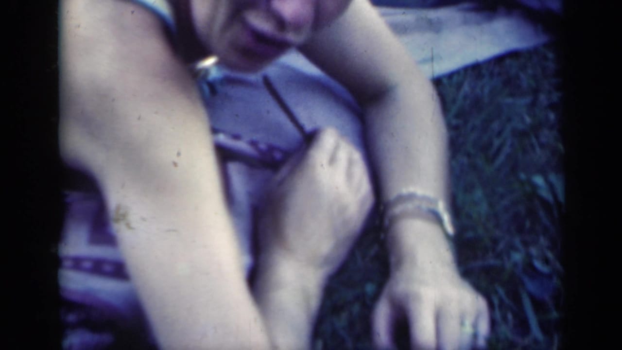 The young woman reclines outdoors on a blanket, with a bracelet, patterned clothing, and grass visible around her.