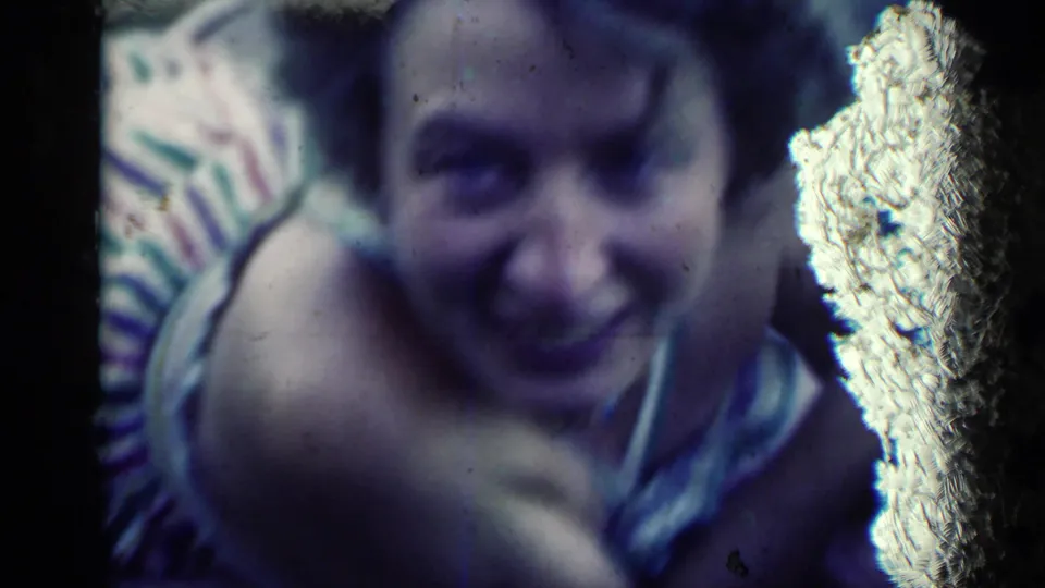 The young woman smiles toward the camera in a soft 8mm close-up, partly obscured by a bright physical-film mark.