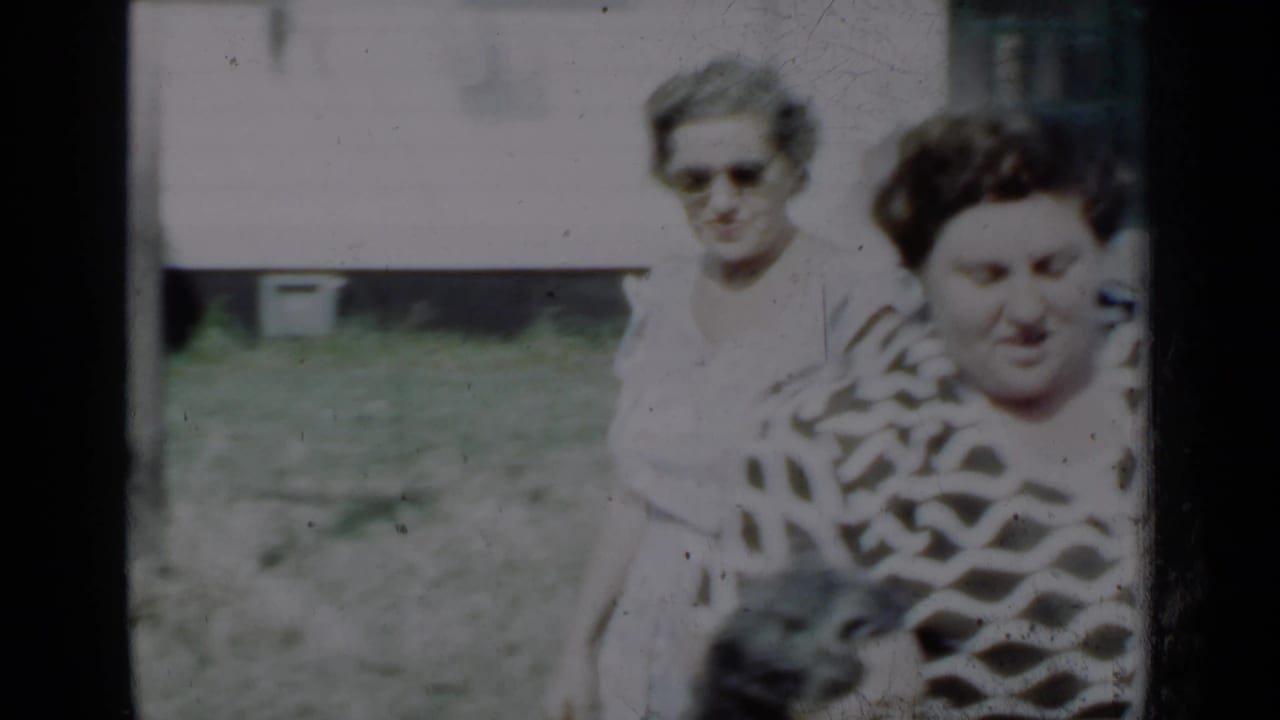 Two adult women in a sunny yard near a house, one in a white dress and one in a patterned top.