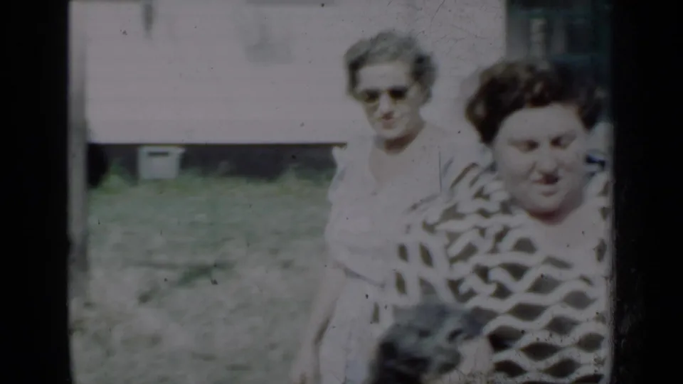 1950 New York home-movie frame showing two adult women outdoors near a house, with one in a white dress and another in a patterned blouse.