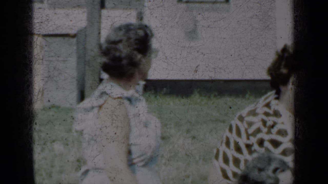 Woman in a white dress seen from behind beside another woman near house siding and grass.