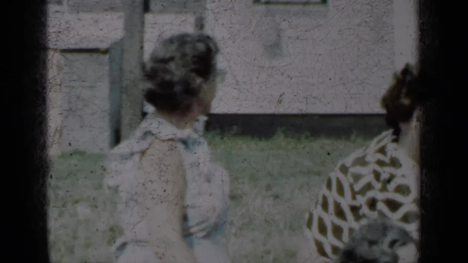Adult woman in a white dress is seen from behind in a New York yard, with another woman partly visible near the house.