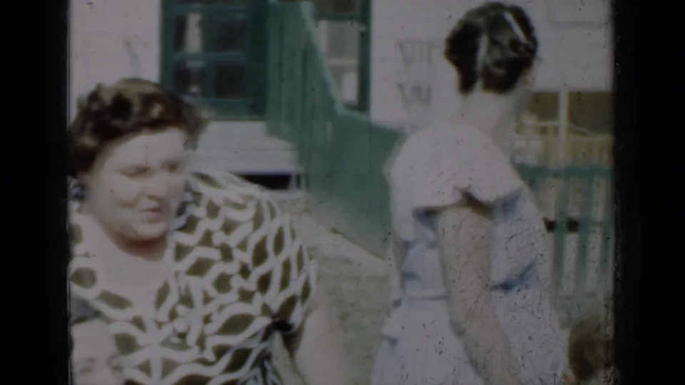 Smiling adult woman in a patterned blouse appears in front of green porch steps, with a woman in a white dress nearby.