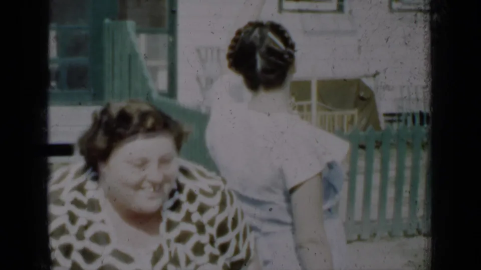 1950 New York home-movie frame with a smiling woman in a patterned blouse and a woman in a white dress near a green porch railing.