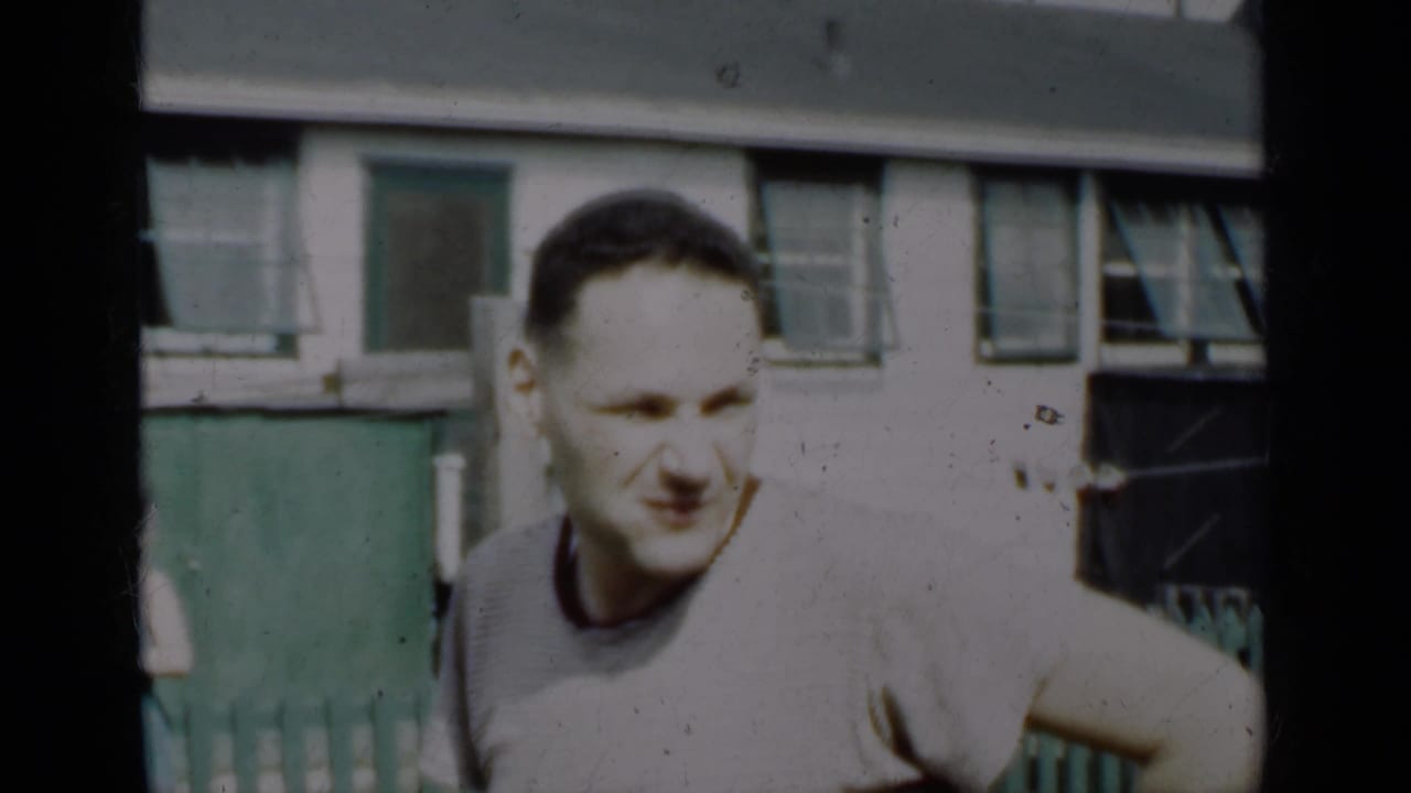 Young man in a light shirt standing by a green fence in bright sun.