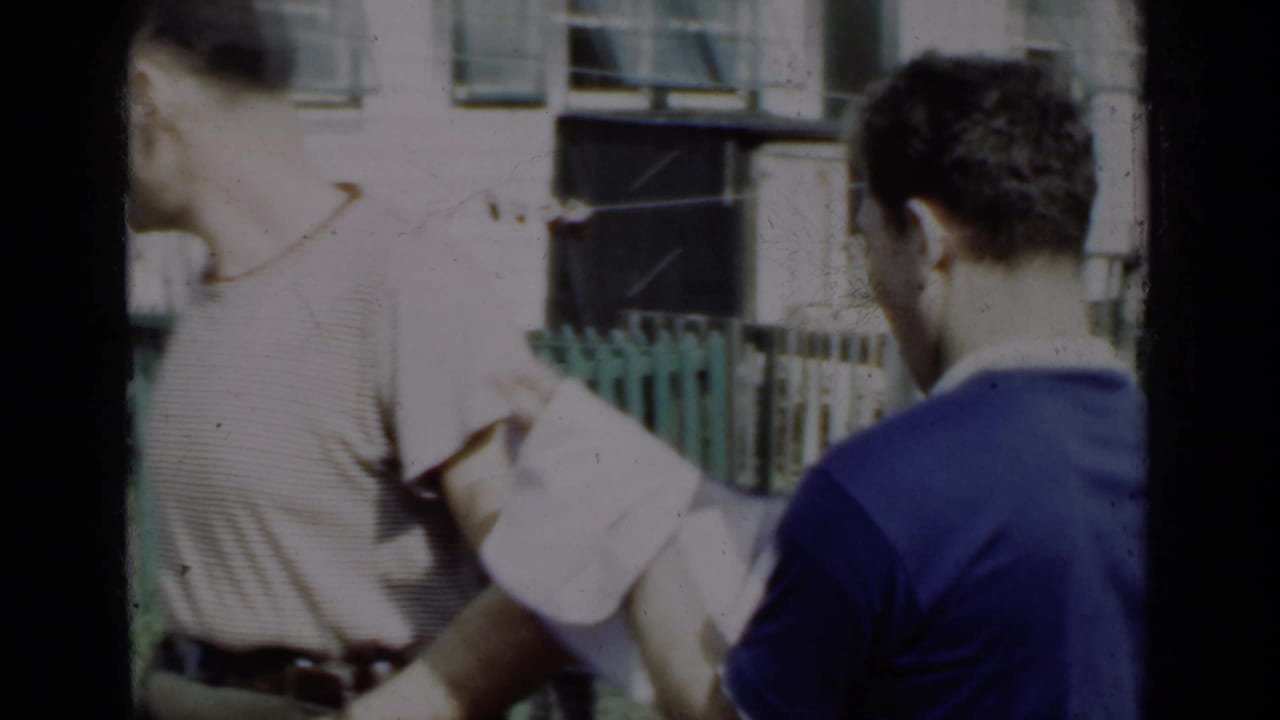 Two men outdoors near a green fence, one partly turned away.