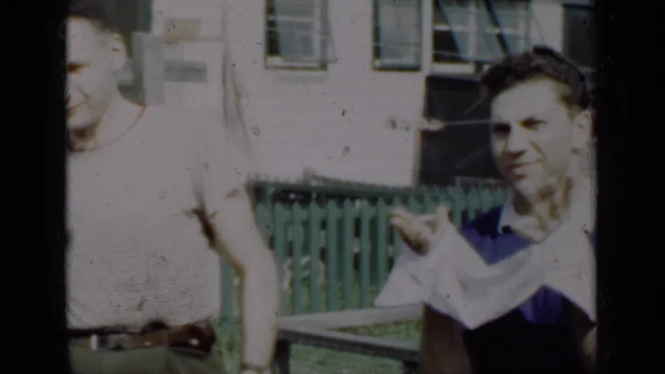 1950 New York home-movie frame showing men beside a green fence, including a man in a blue shirt gesturing with a light cloth.