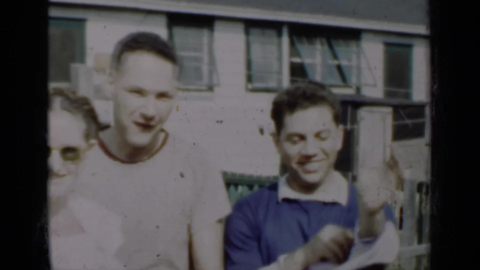 1950 New York 8mm frame of three adults near a green picket fence, with two men facing the camera and smiling or gesturing.