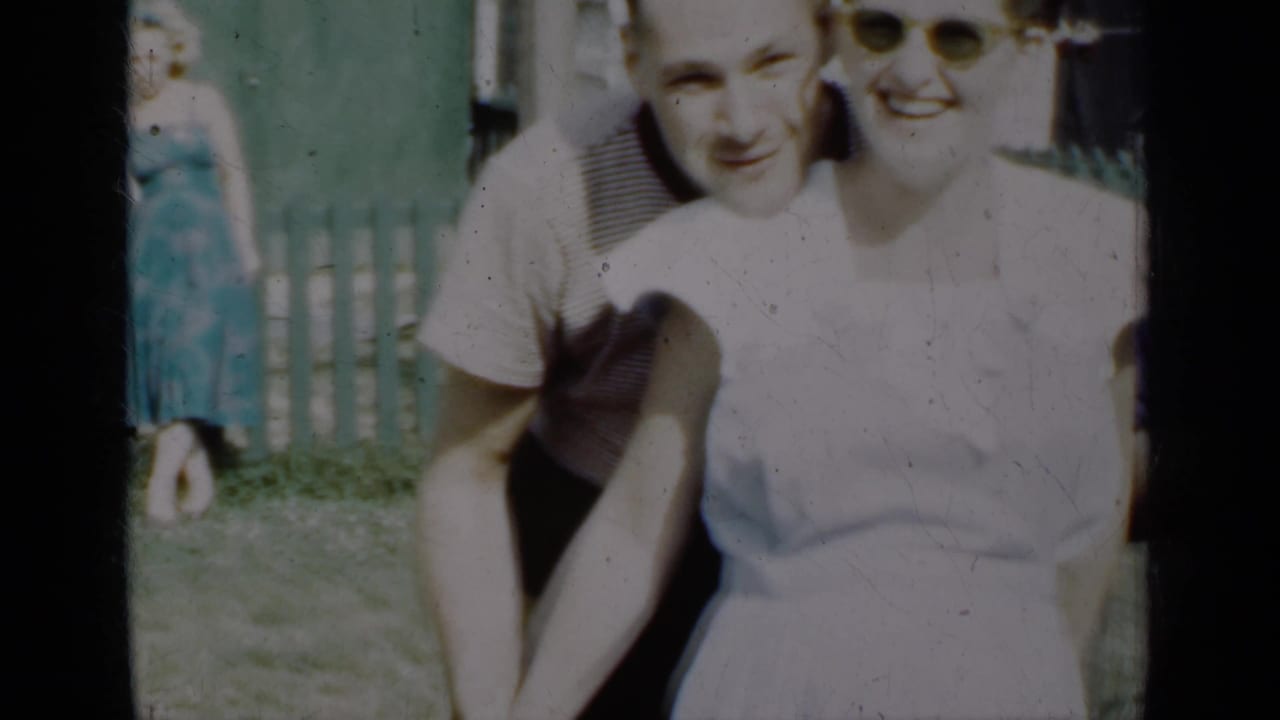 A man leans behind a smiling woman outdoors by a fence.