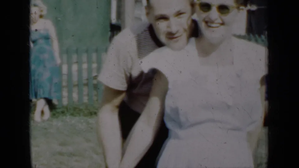 A man leans close behind a smiling woman in a white dress during a 1950 New York outdoor home-movie moment.
