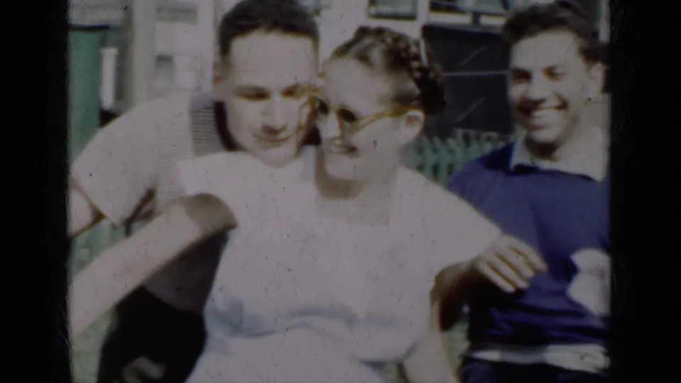 Two men and a woman lean together in 1950 New York home-movie footage, laughing in front of a house and fence.
