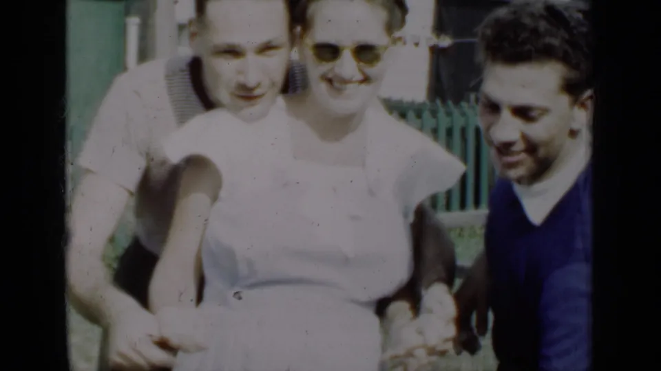 Three young adults in 1950 New York footage smile and gesture in a sunny residential yard with a fence behind them.