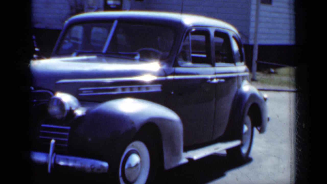 Dark rounded sedan parked on a sunlit street beside a light-sided building.