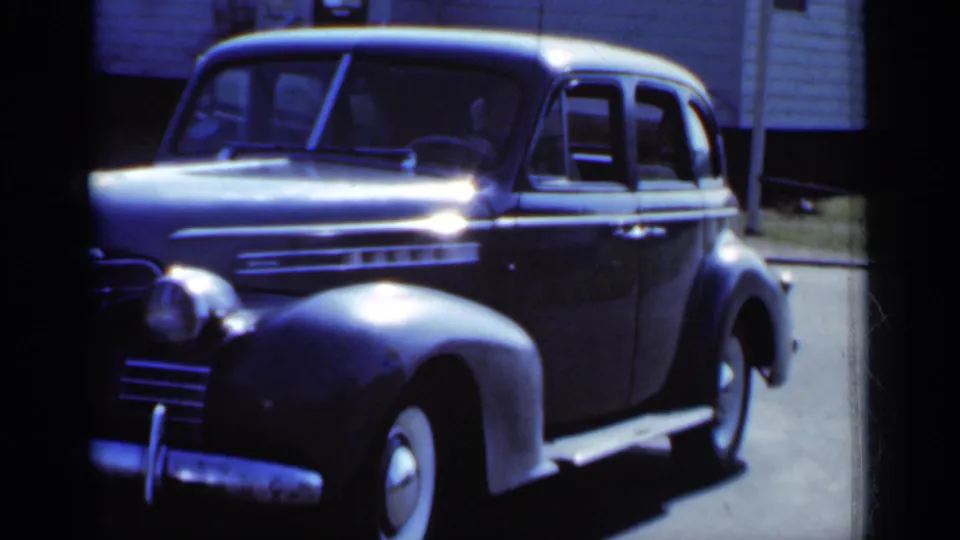 1950 New York frame of a dark parked sedan, its hood and chrome details catching daylight near a residential exterior.
