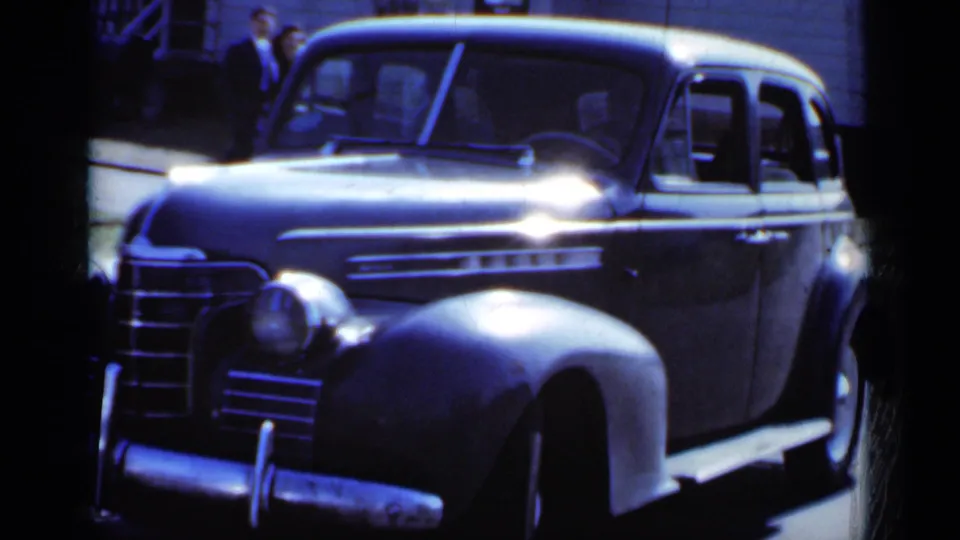 1950 New York home-movie frame showing the parked dark sedan closer to camera, with a few distant people at the left background.