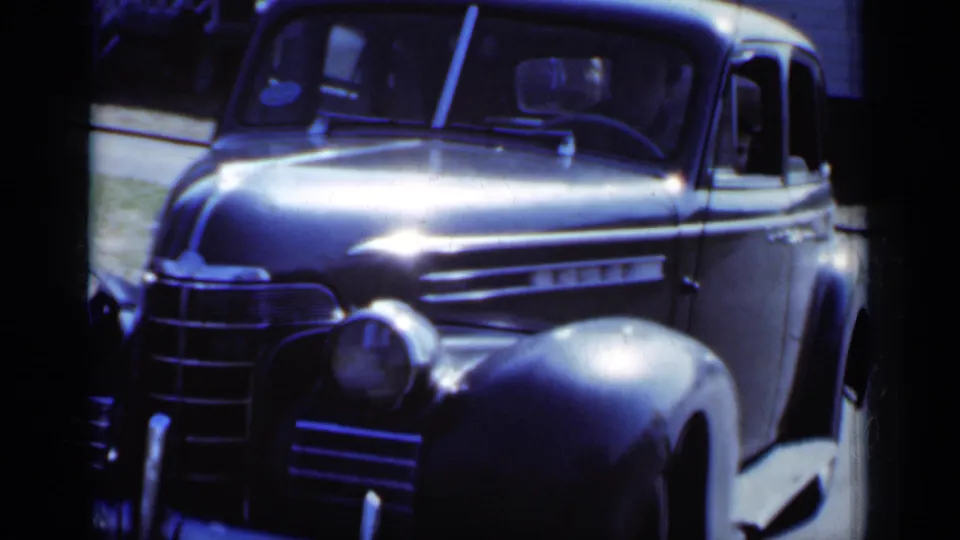 1950 New York frame showing a dark mid-century sedan from a front angle, with chrome grille, rounded fender, and side windows visible.