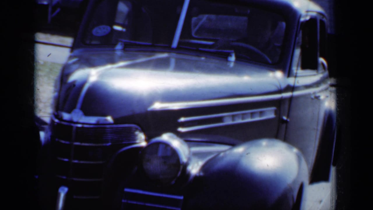 Front of a rounded vintage sedan with chrome grille and fender in blue-tinted 8mm footage.