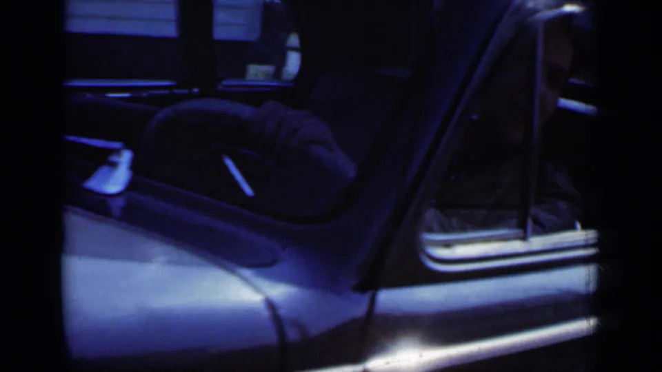 Driver-side view of the vintage sedan, with the door open, steering wheel visible, and the dark interior partly framed by handheld 8mm footage.