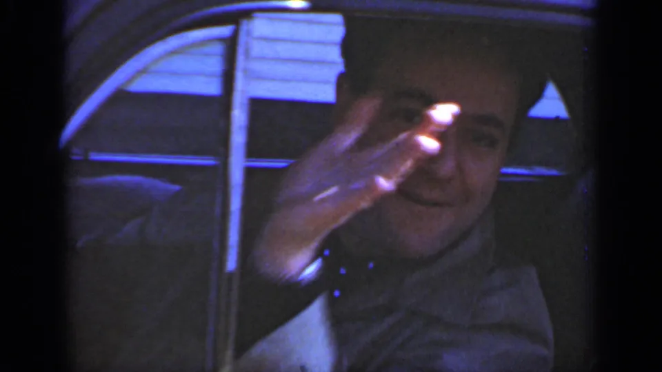 Adult male driver in a New York 1950s home movie smiles from inside the sedan and raises his hand in a casual greeting toward the camera.