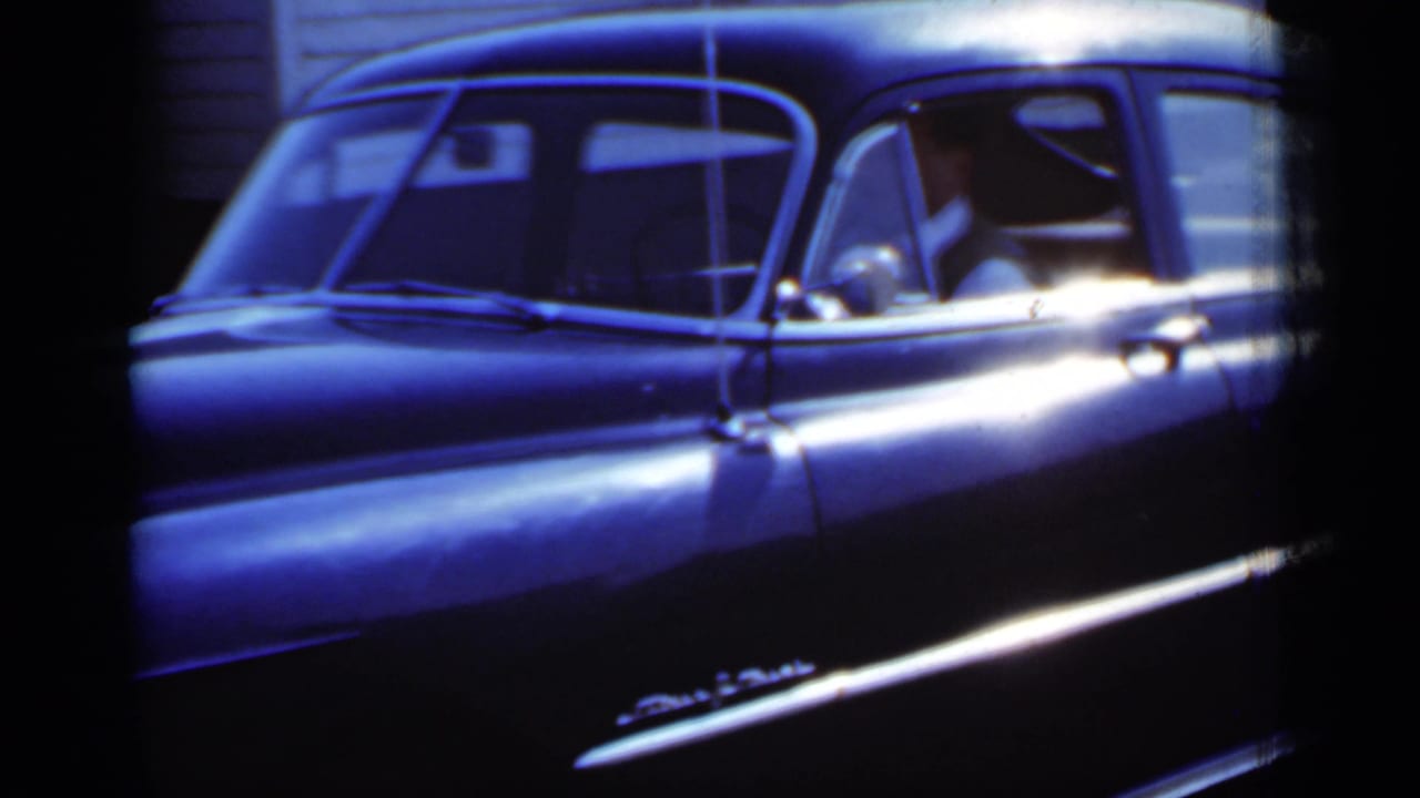 Side view of a dark vintage sedan with a seated driver and bright reflections on the body.