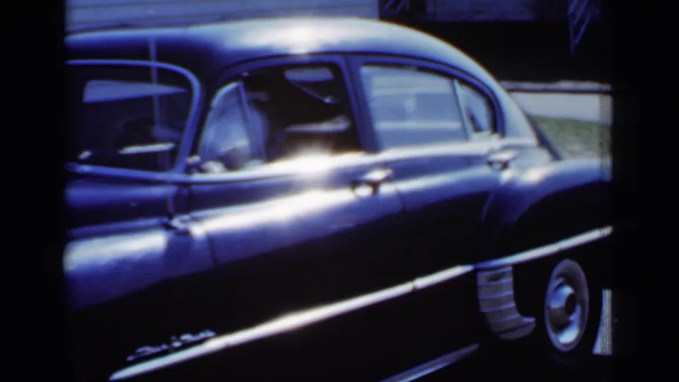1950 New York 8mm frame showing a dark chrome-trimmed sedan in sunlight, with the driver area and rear wheel visible.