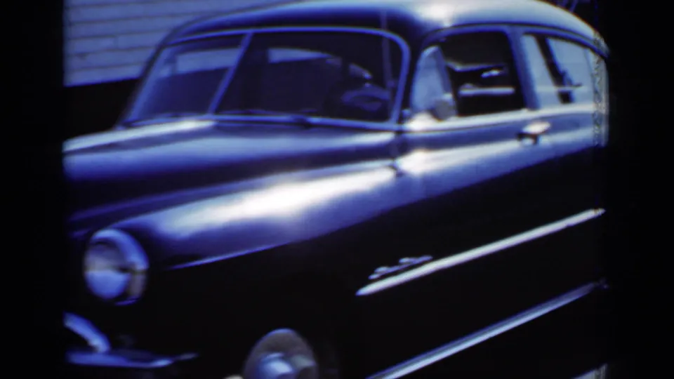 A black sedan with chrome side trim appears close to the camera in 1950 New York home-movie footage, with residential siding behind it.