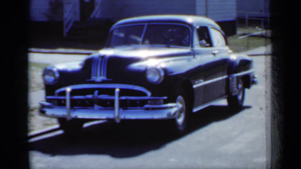 The black sedan is shown from the front in a New York residential setting, with chrome grille, bumper guards, and windshield visible.