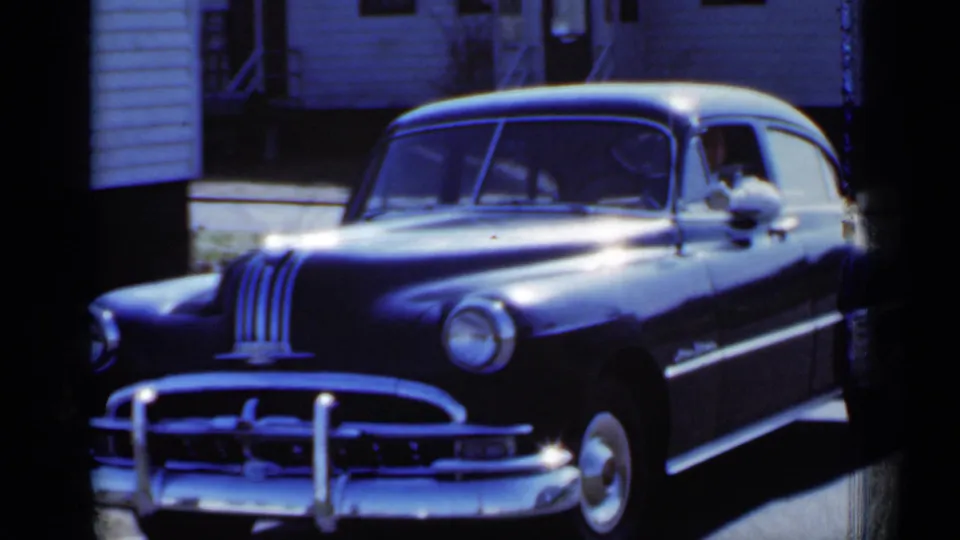 A front three-quarter view shows the black sedan in 1950 New York, with chrome details and a passenger visible near the side window.