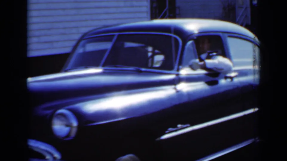 A passenger in a light shirt waves from the open window of the black sedan in 1950 New York home-movie footage.