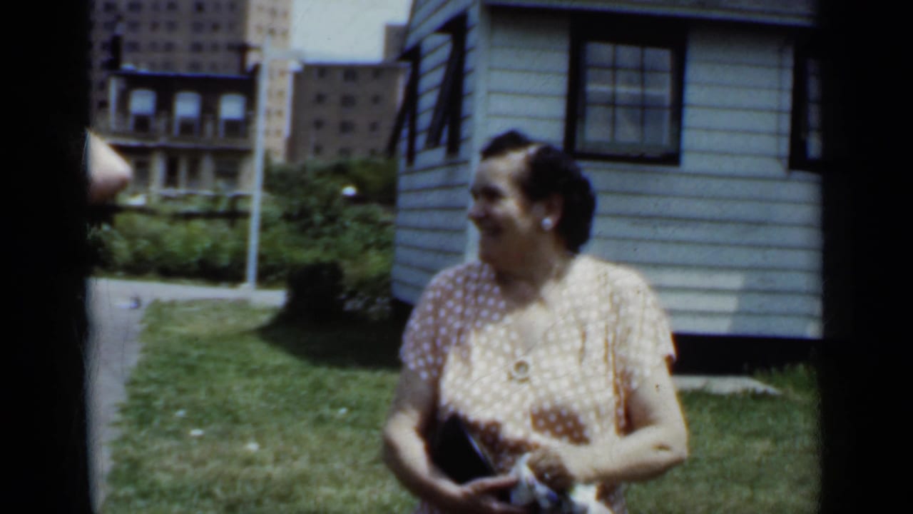 Woman in a polka-dot dress standing in a grassy urban yard near a light-colored house.