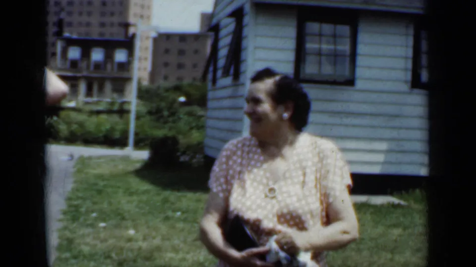 In 1950 New York home-movie footage, a woman in a polka-dot dress stands smiling in a grassy yard with city buildings behind her.