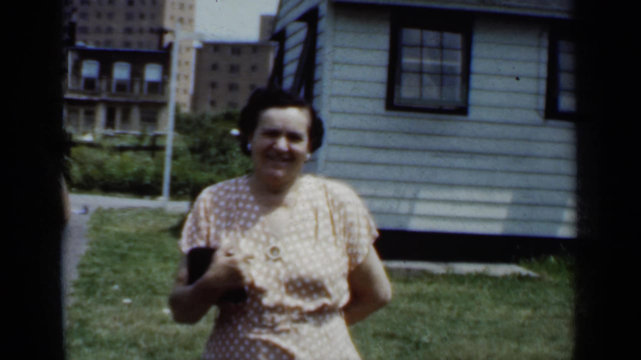 The woman faces the camera in the yard, holding a dark object, with a house wall and high-rise buildings in the background.