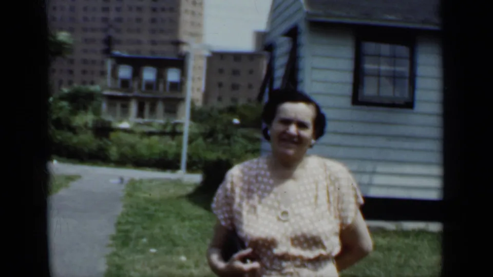 A wider home-movie frame shows the woman in the yard beside a light-colored house, with paths, shrubs, and city buildings behind.