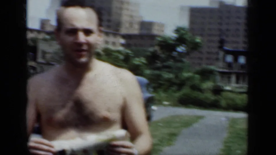 A shirtless adult man stands in the same urban yard setting, holding a light cloth with apartment buildings visible in the background.