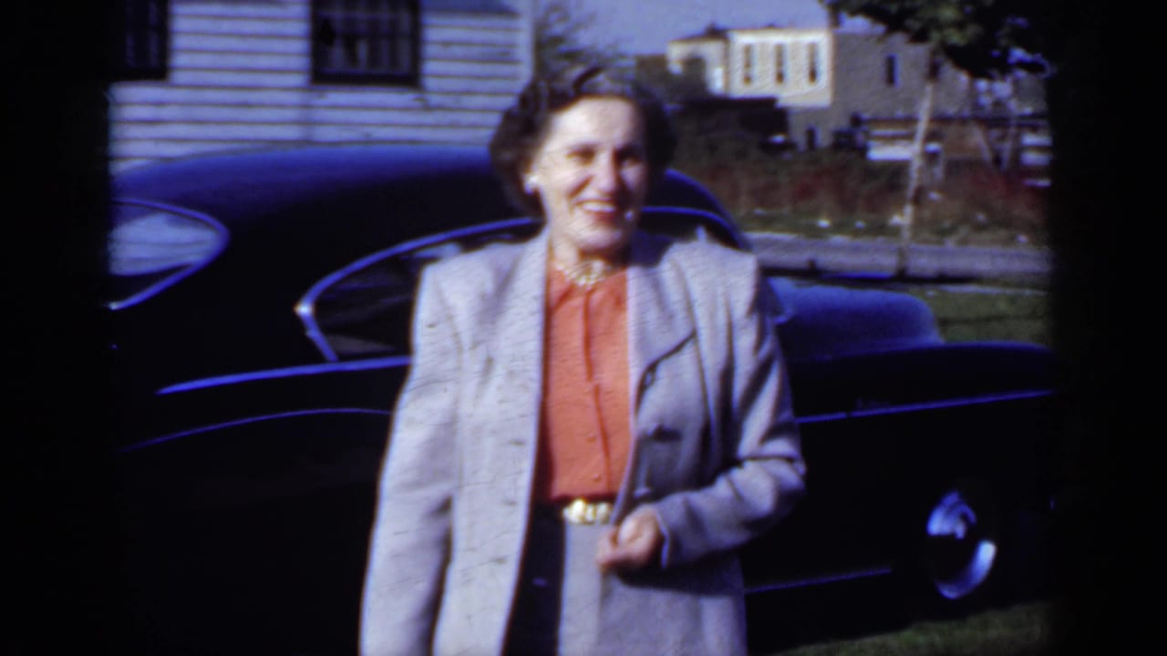 In 1950 New York home-movie footage, a woman in a grey suit and coral blouse smiles beside a dark car in a sunny residential setting.