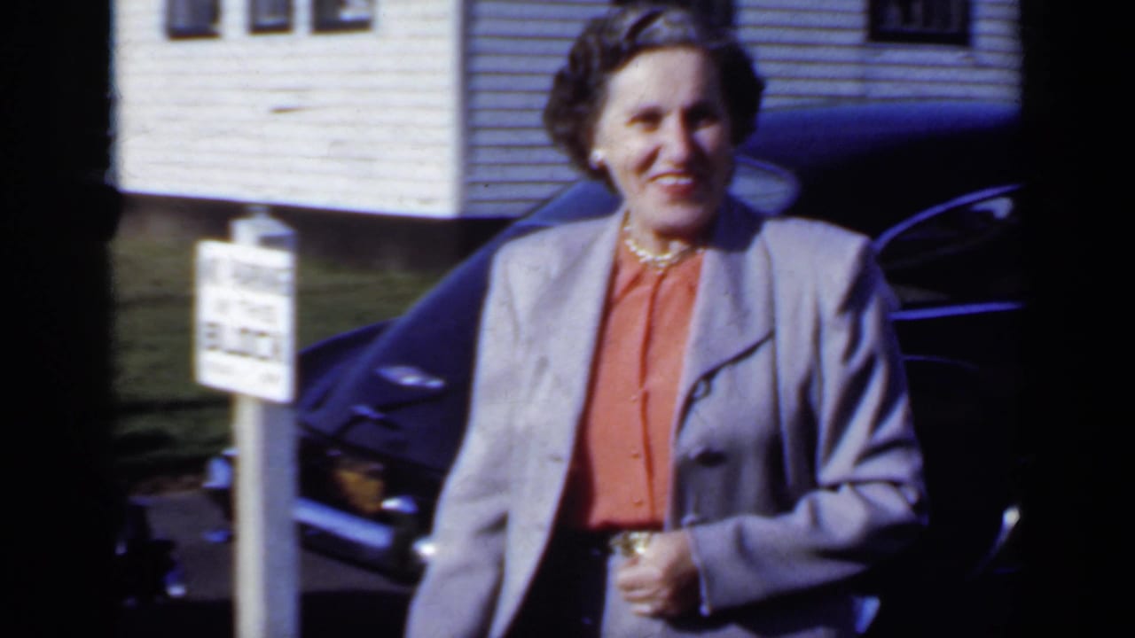 Woman in a grey suit walks past a dark car with a white house behind her.