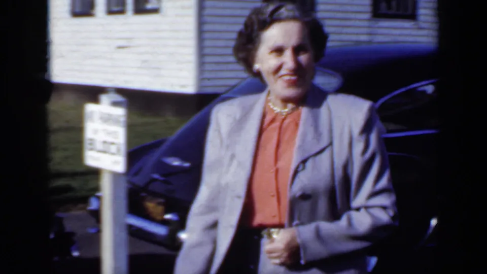 The woman walks past a dark mid-century car in bright sunlight, with white clapboard houses visible behind her in New York footage from 1950.