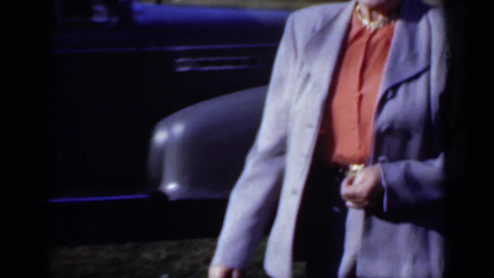 A closer, partly cropped frame shows the woman's grey suit, coral blouse, necklace, and belt area as she moves past the dark car.